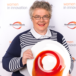 Kathy holds artistically painted plate in front of a display board decorated with Women In Innovation logos